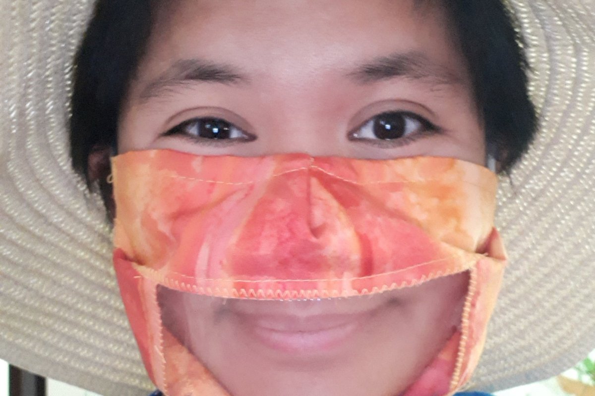Woman wearing an orange fabric mask with plastic windown showing her smile, and wearing a straw floppy hat.