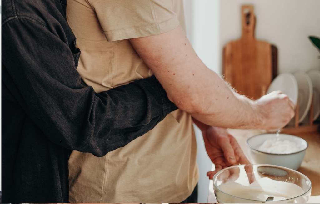 Close up of a couple mixing baking ingredients. One person hugging the other from the back. 