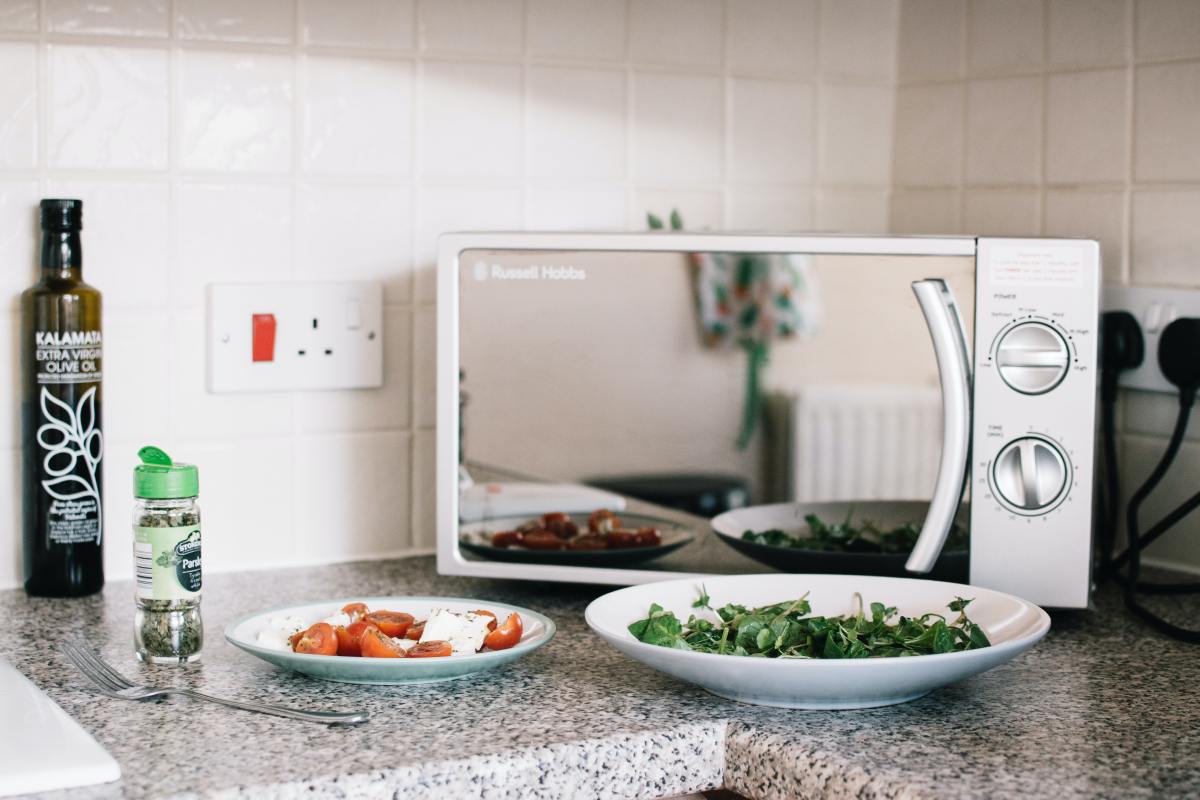 Kitchen counter with a microwave, sauce with tomatoes, a plate with greens and bottle of spices and olive oil