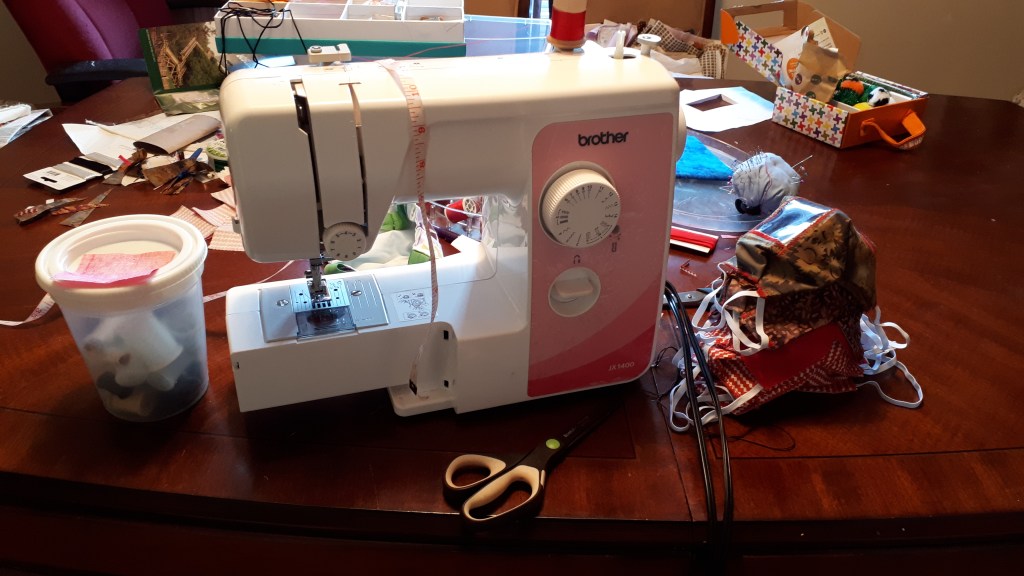 Sewing machine on a wood table with sewing supplies around it, and a pile of completed fabric face masks beside the machine.