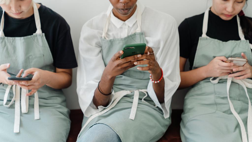 Three people wearing uniforms and aprons sitting beside each other browsing on their own smartphones