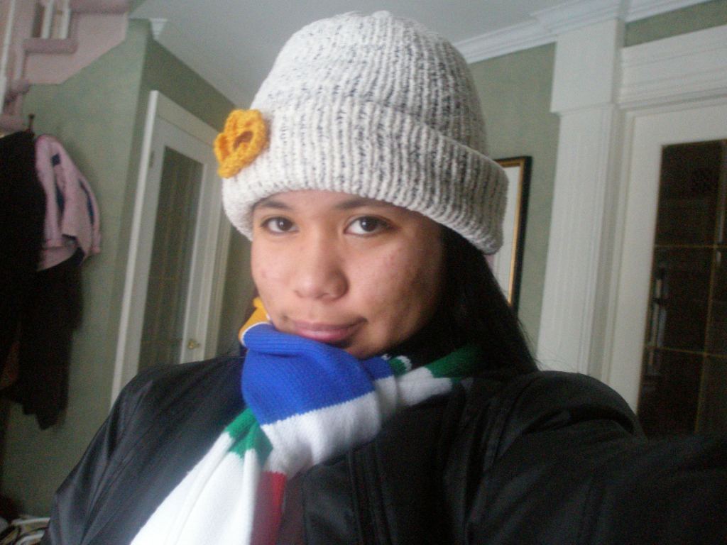 Woman wearing a black winter jacket, multi color scarf and white knitted hat with a yellow knitted flower.