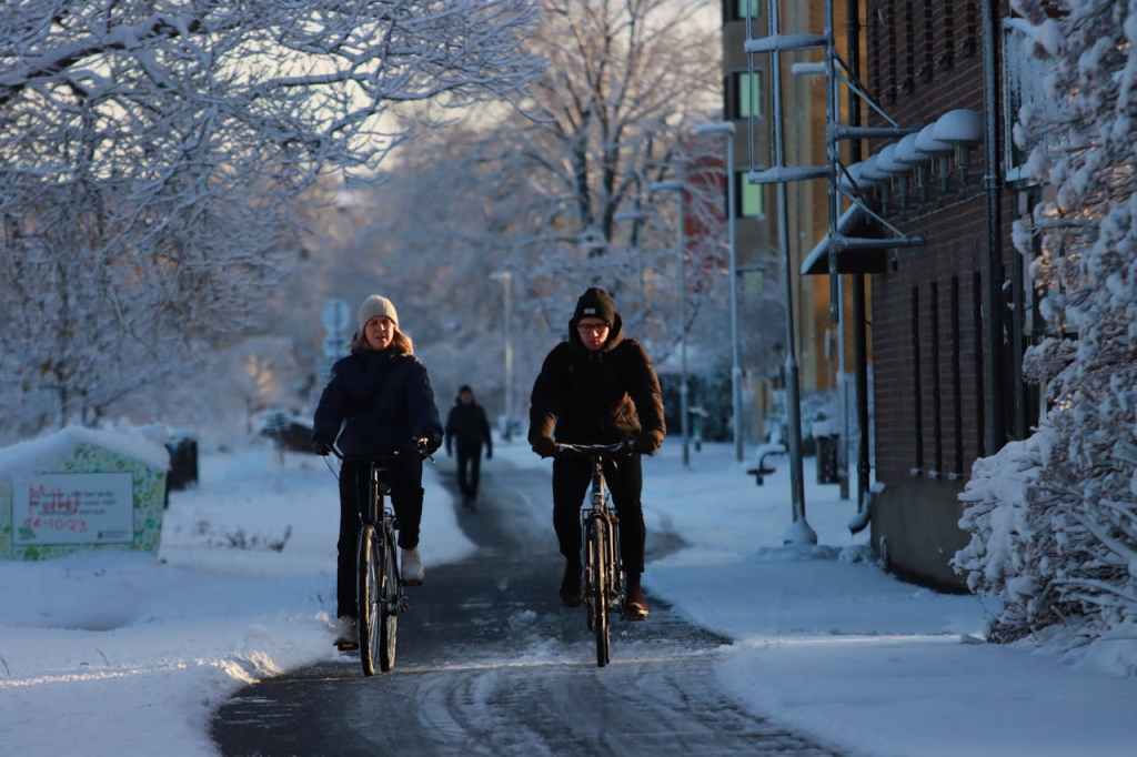 Two people biking on a bike path in winter. Everything, except for the actual bike path, is covered in snow.